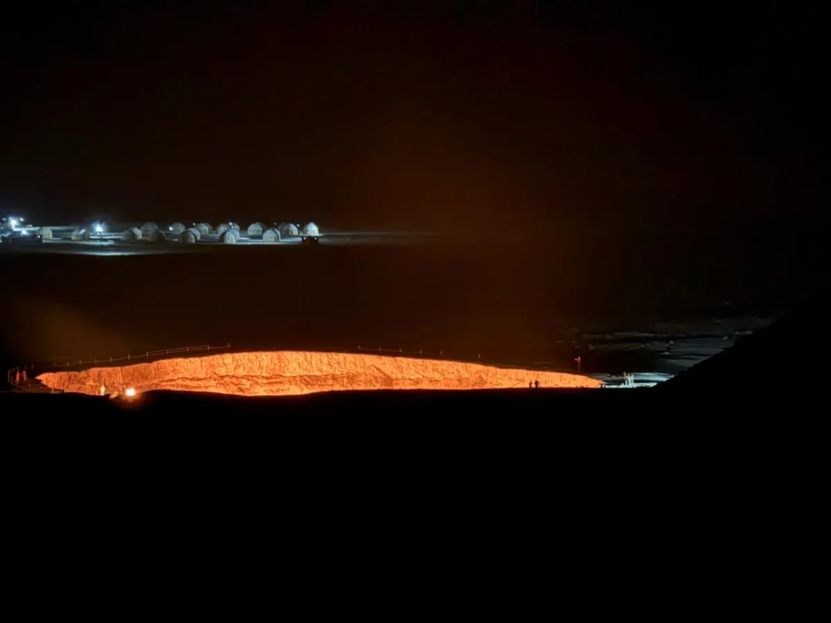 The blazing Darvaza gas crater at night and a glowing yurt camp against the backdrop of the dark desert in Turkmenistan