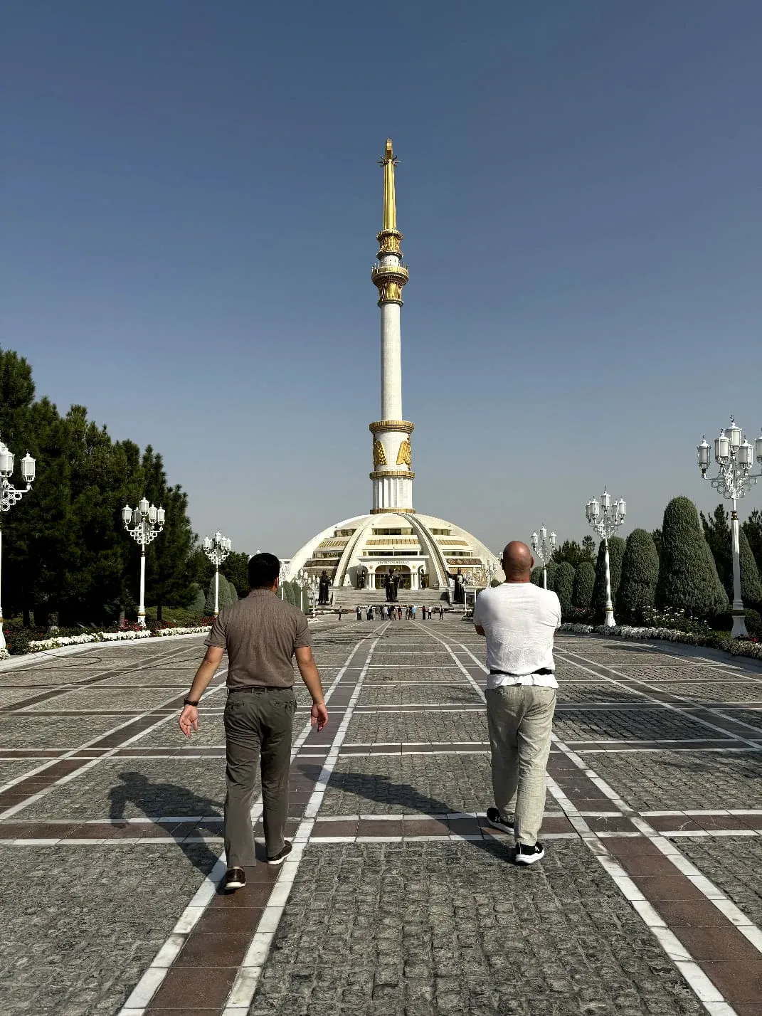 Two tourists walk along a wide paved avenue towards the Independence Monument in Ashgabat, Turkmenistan