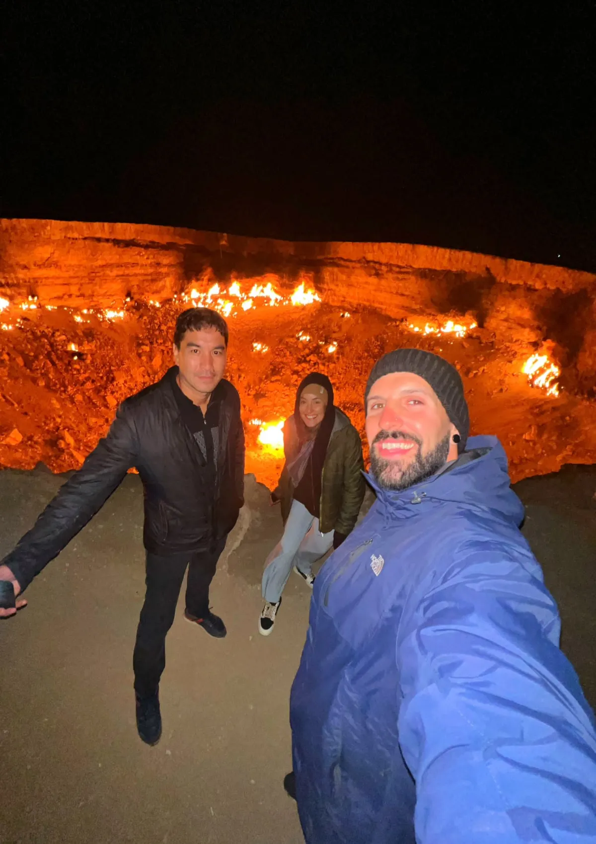 Un groupe de trois touristes souriants prend un selfie nocturne au bord du cratère gazier brûlant de Darvaza