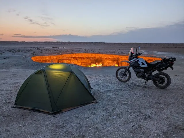Tente verte illuminée de l'intérieur et moto de voyage au bord du cratère gazier en feu de Darvaza au crépuscule