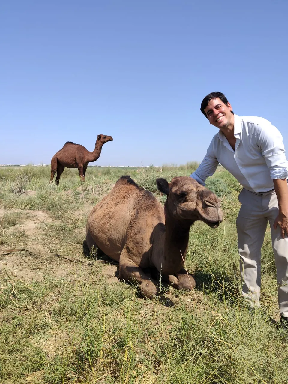 A smiling tourist stands next to a reclining camel on the steppe of Turkmenistan under a clear blue sky.