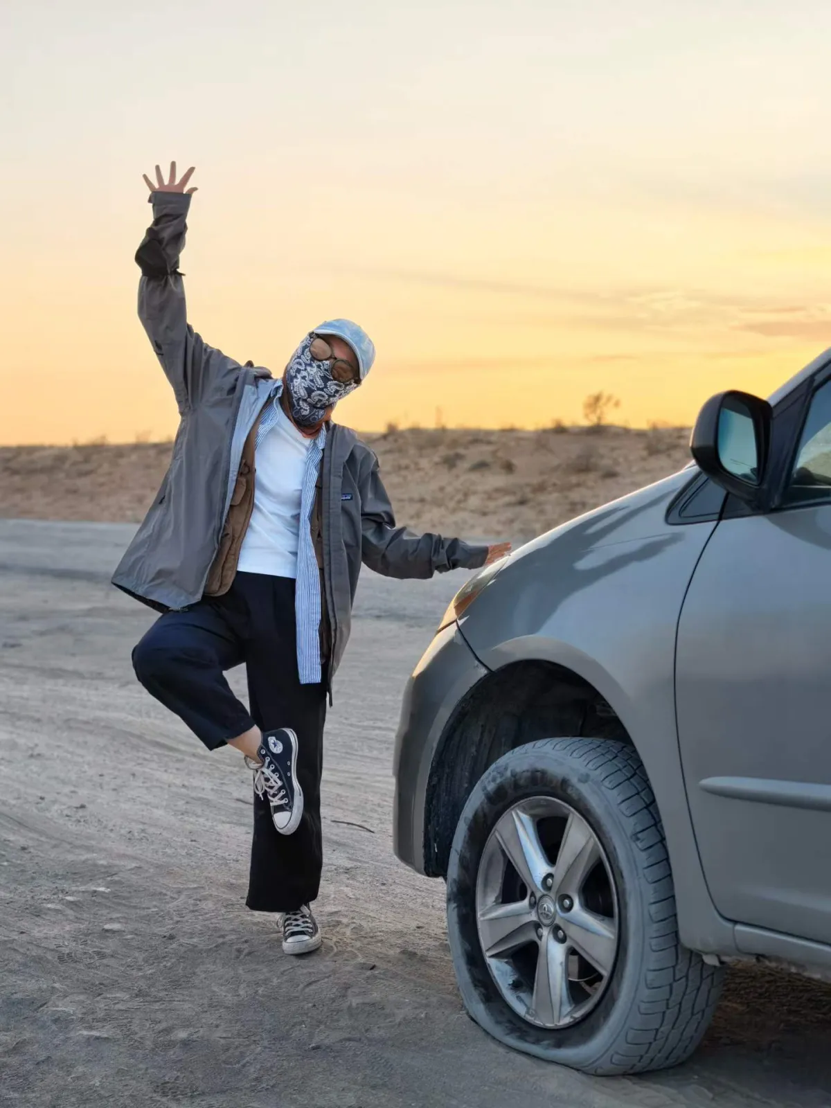 A traveler playfully posing on one leg next to an off-road vehicle in the Darvaza desert at sunset