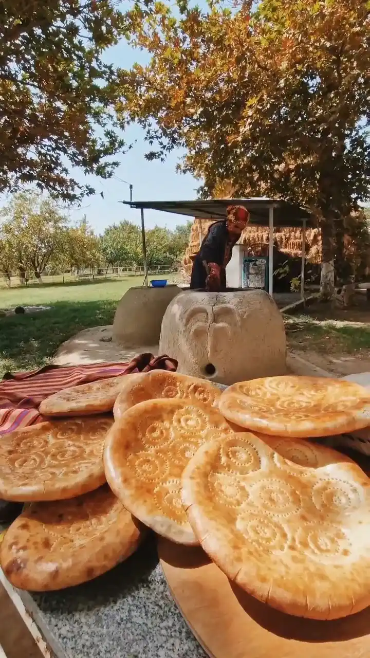 Una donna turkmena cuoce il pane tradizionale in un tandoor di argilla; in primo piano, pagnotte calde rotonde con motivi decorativi — autentica vita di villaggio in Turkmenistan.