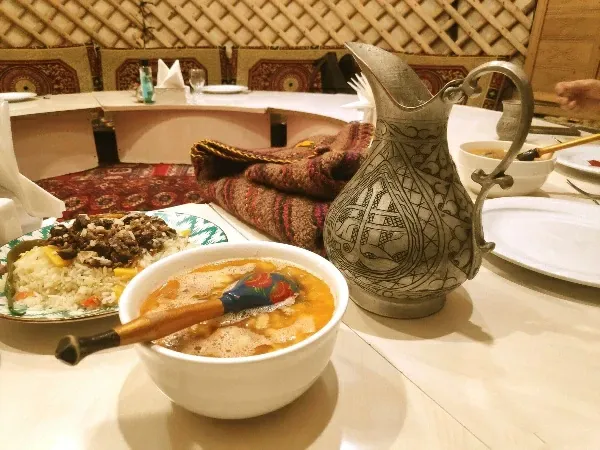 Traditional Turkmen dograma soup with a wooden spoon, a plate of plov, and a metal jug of ayran on a table inside a national yurt