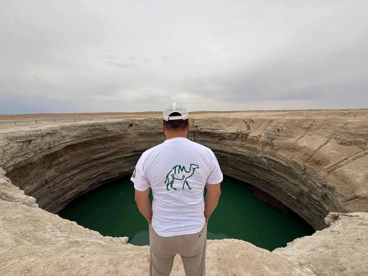 A person in a white t-shirt with a camel logo stands on the edge of a huge circular crater with turquoise water in the Turkmenistan desert